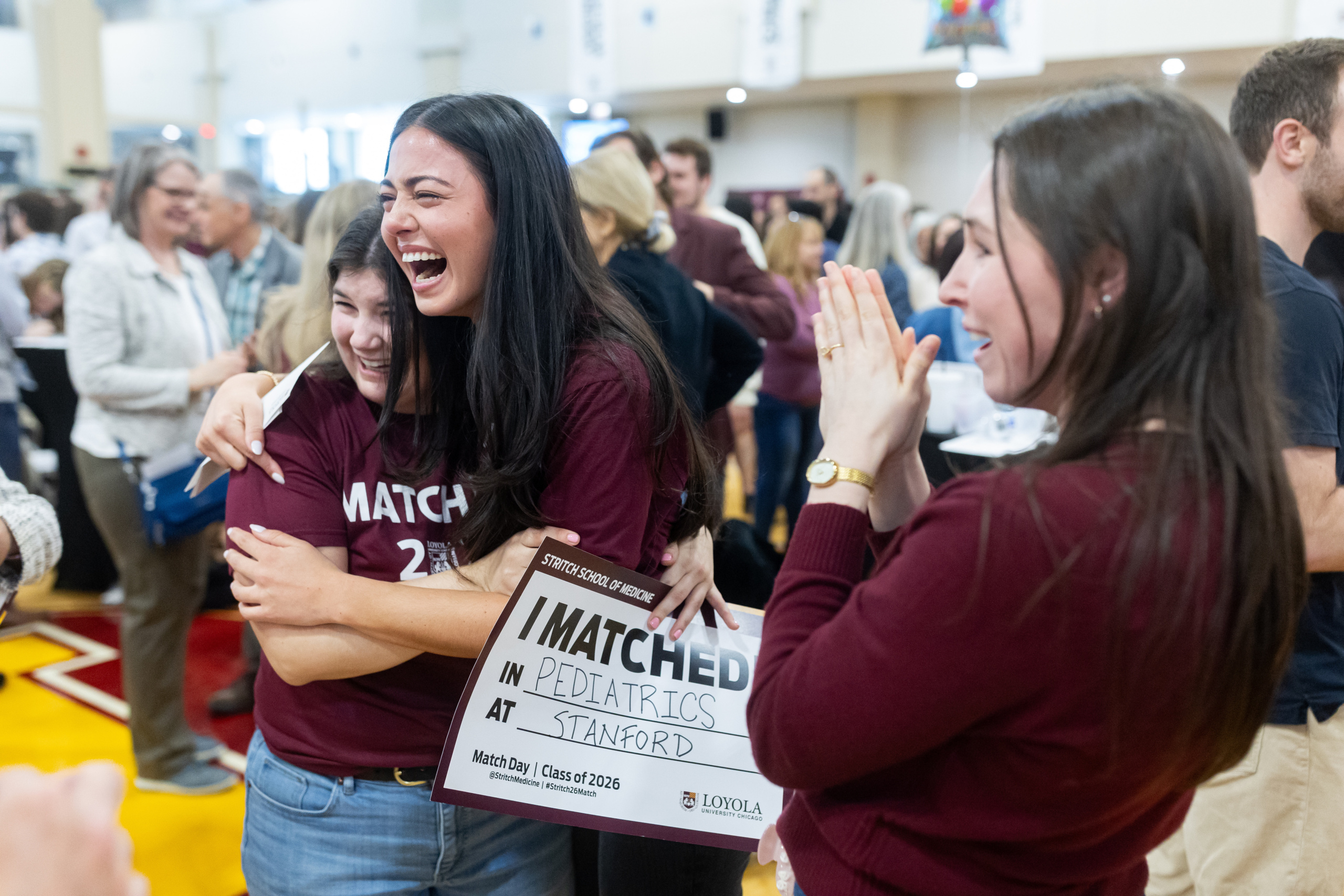 Three women Loyola University Chicago students wearing maroon T-shirts embrace, clap, and cheer in a gymnasium, with one woman holding a sign reading "I MATCHED"