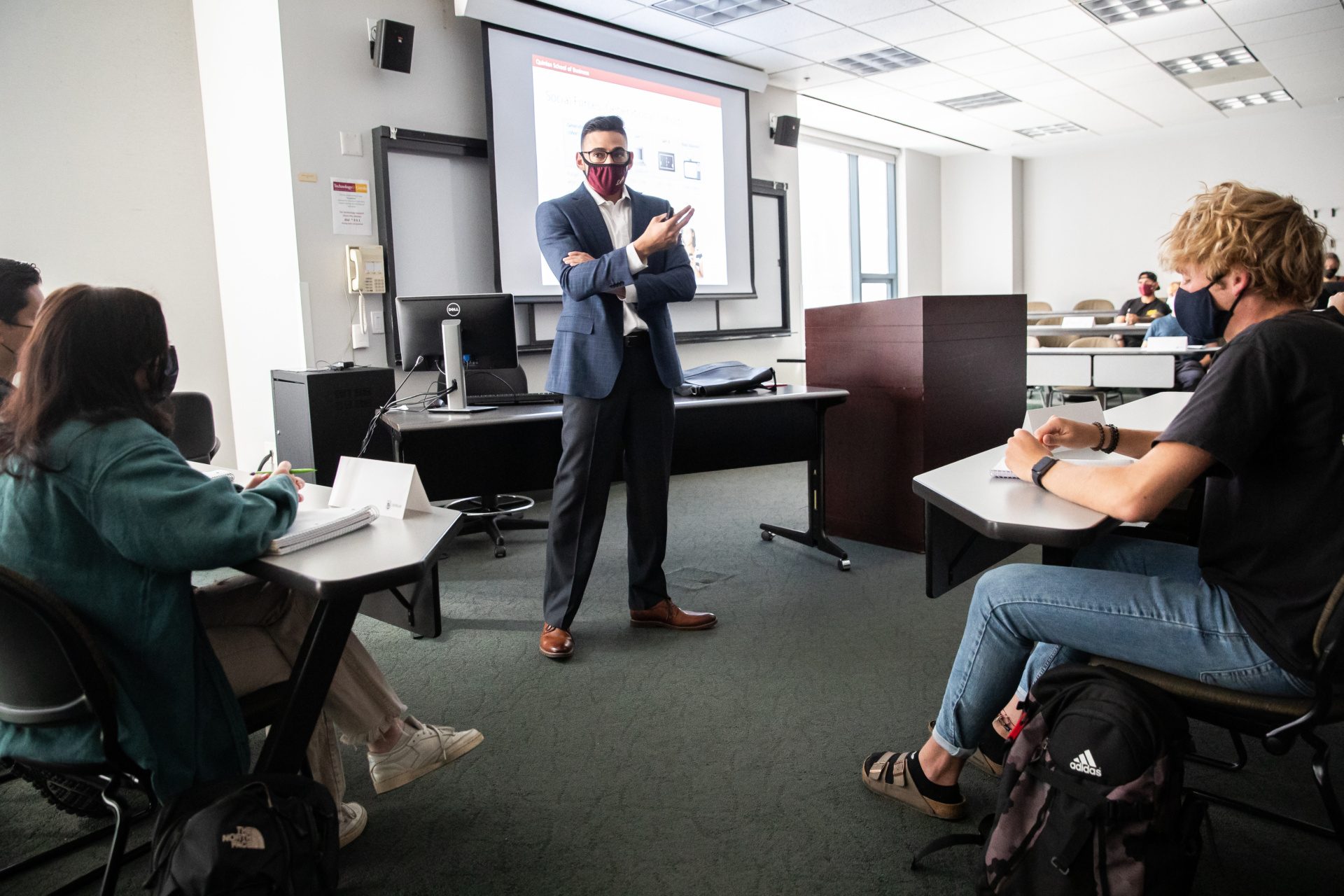 A man wearing a suit and a Loyola University Chicago mask crosses his arms in a classroom while standing in front of a Powerpoint presentation