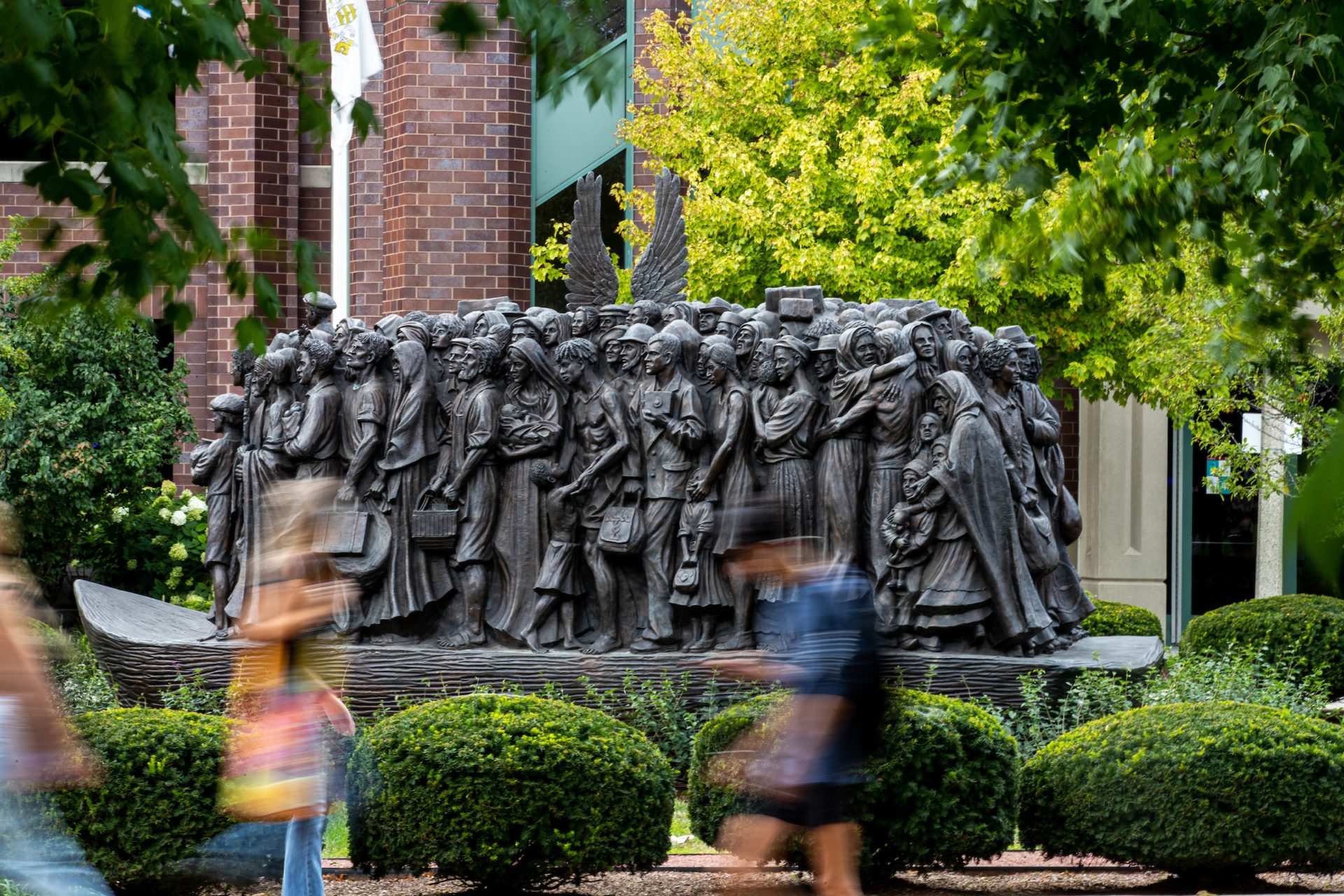 Blurred students pass a large bronze statue depicting a large group of migrants with a pair of angel wings behind them