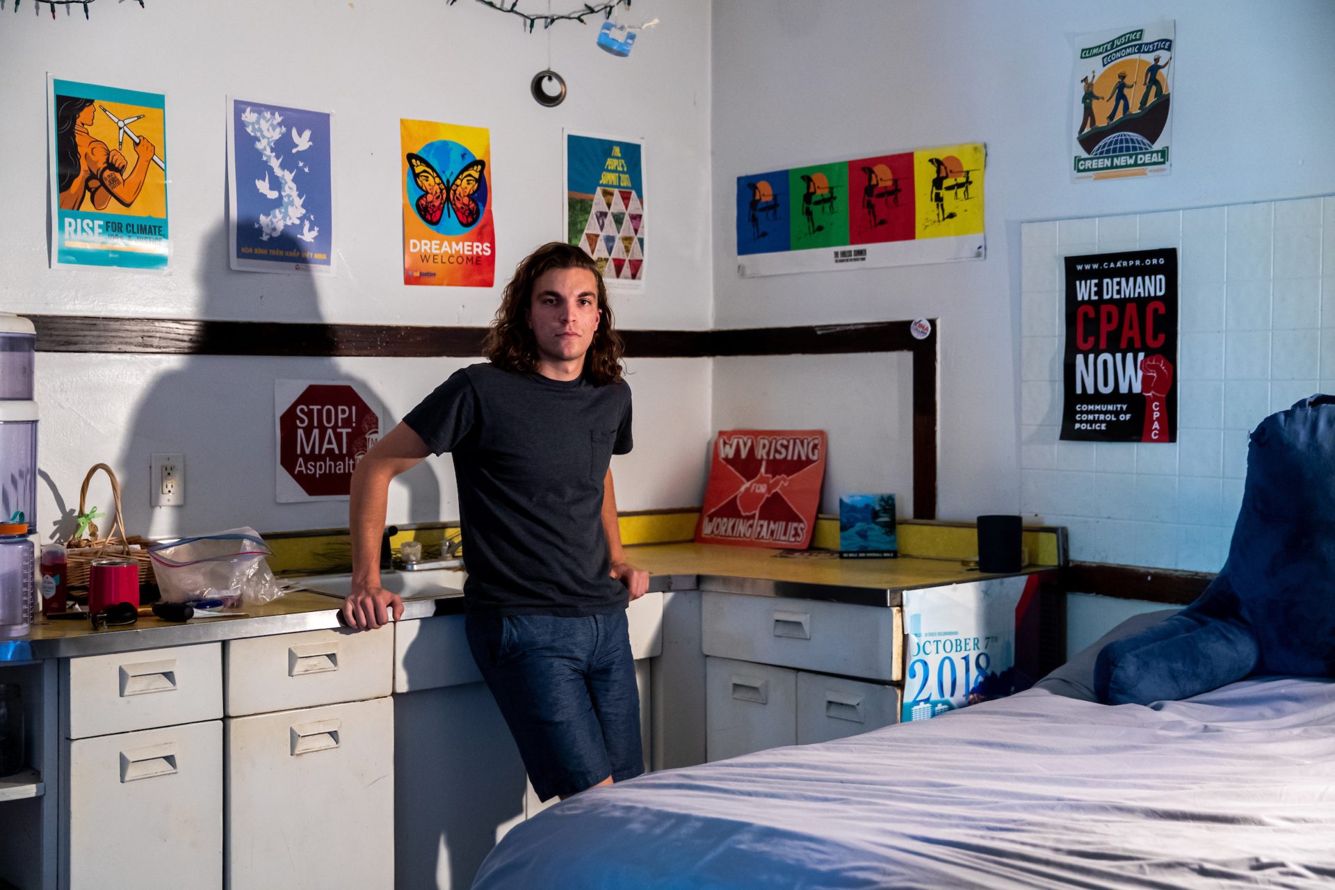 A man with shoulder-length hair leans against a counter in a small apartment with colorful posters on the walls