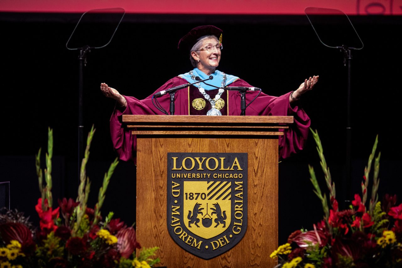 A woman wearing maroon graduation regalia stands behind a wooden podium with the Loyola University Chicago shield