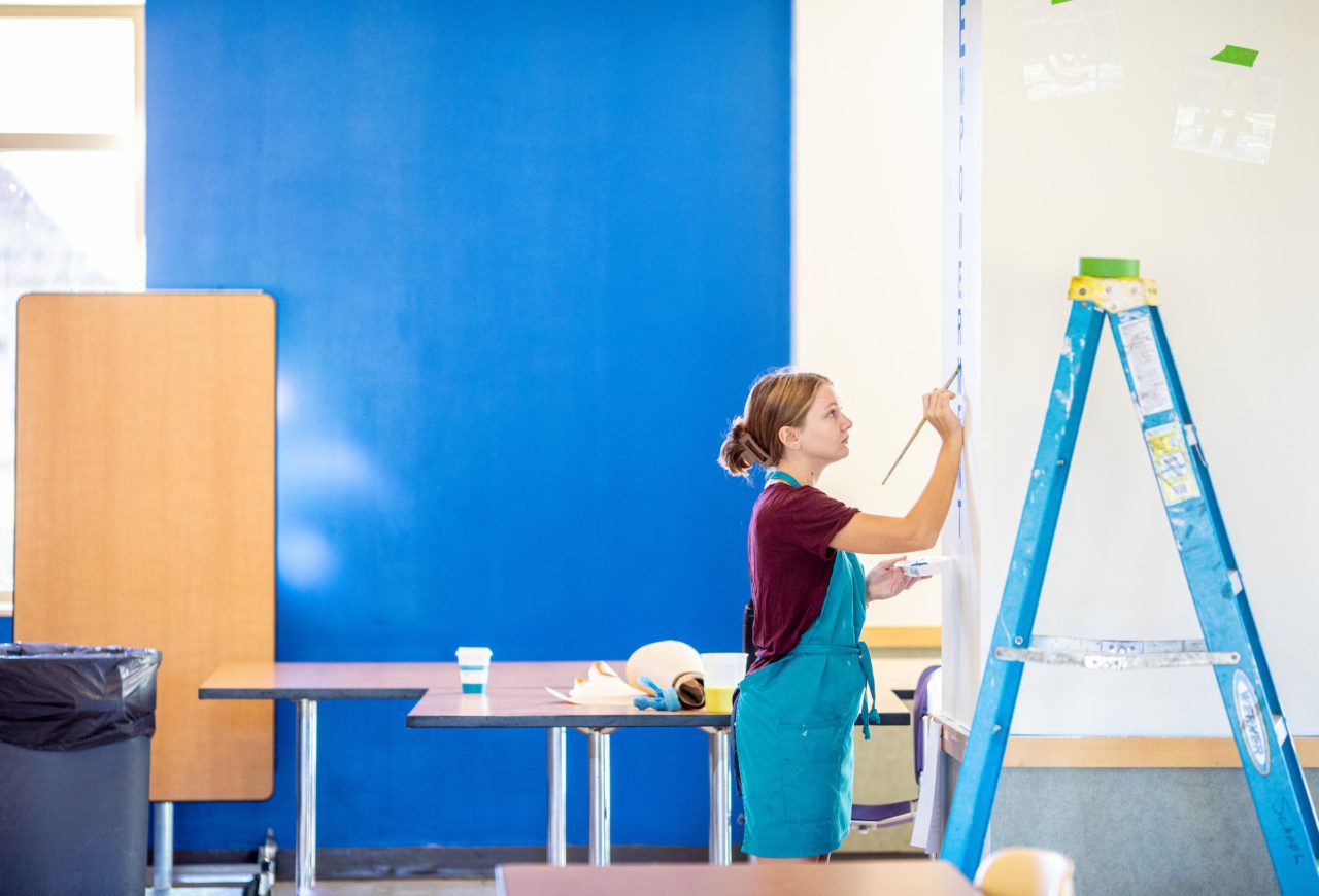 A Loyola University Chicago student wearing a blue apron stands next to a ladder as she paints a wall