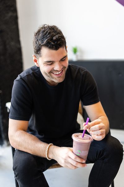 A man wearing a black T-shirt sits in a chair and looks down at a smoothie in his hands