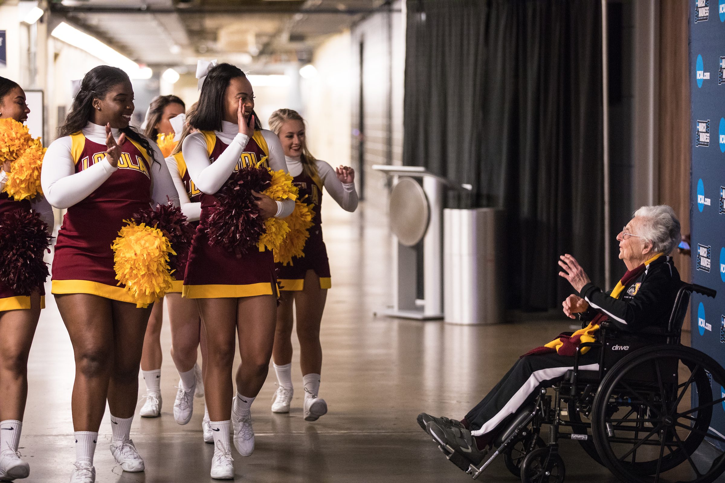 An older woman in a wheelchair waves at a group of women in Loyola University Chicago cheerleading uniforms at a sports stadium