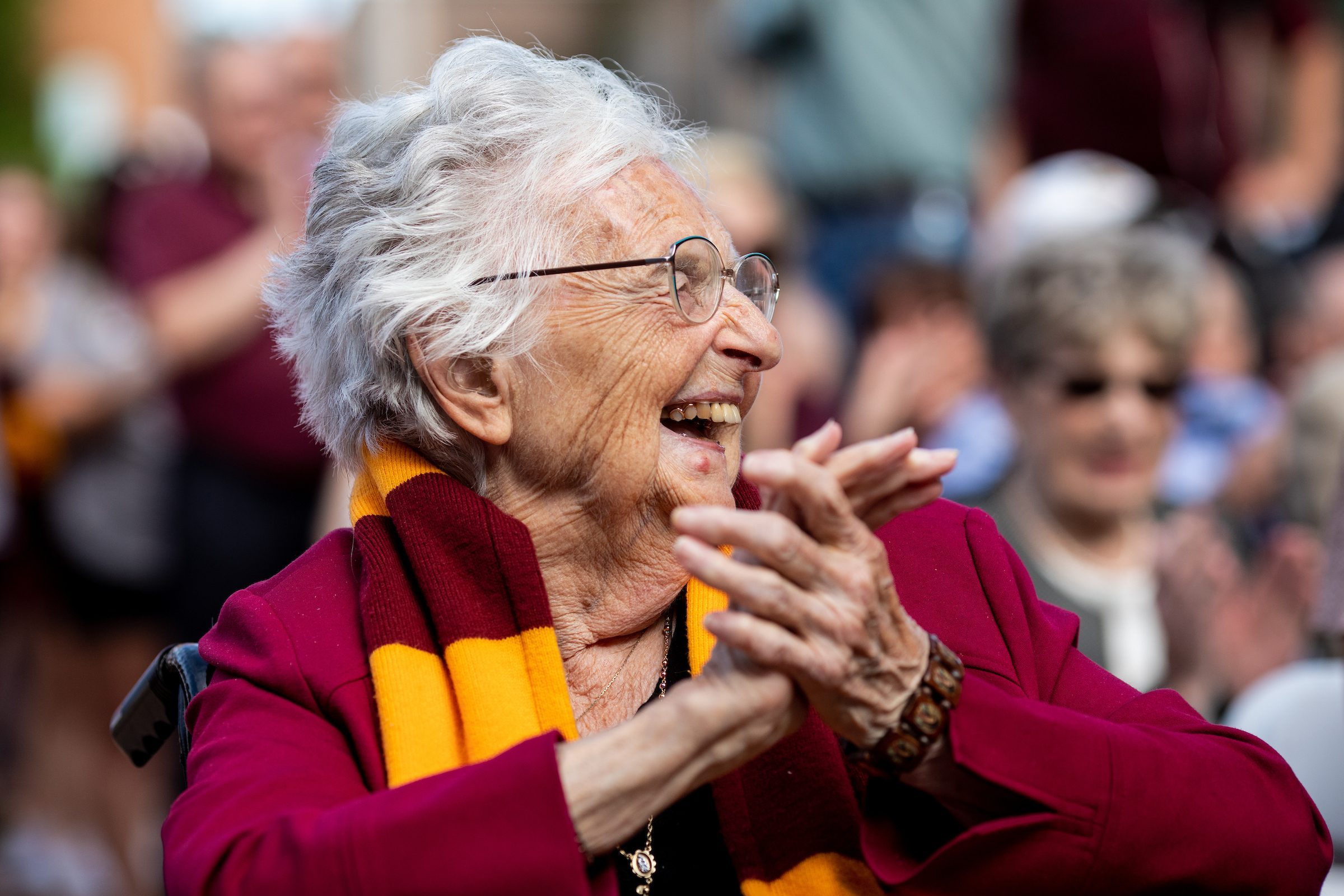 Sister Jean Dolores-Schmidt, BVM, attends a ribbon cutting ceremony for the newly completed Alfie Norville Practice Facility on Sunday, August 25, 2019. (Photo: Lukas Keapproth) An older woman wearing a maroon shirt and Loyola University Chicago scarf smiles and claps with members of an audience in the background