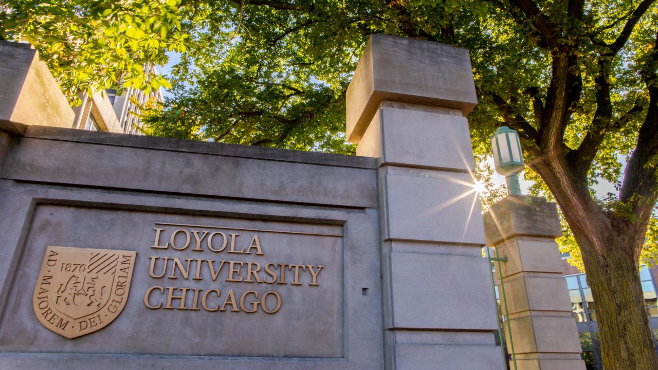 The sun shines through trees behind a campus sign of Loyola University Chicago.