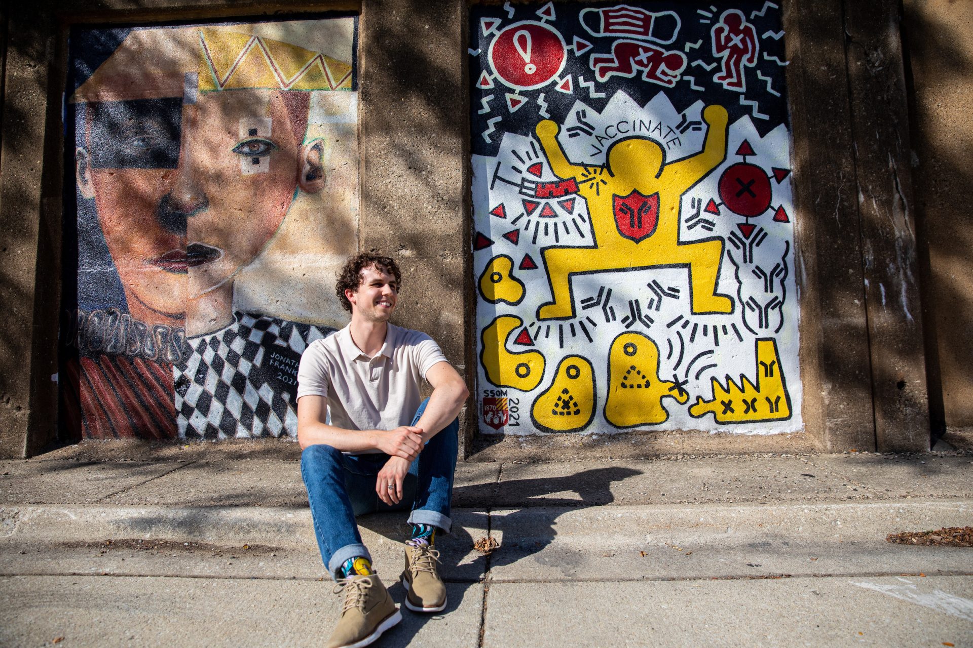 A man sits outdoors in front of a mural depicting a human getting vaccinated and fighting images of illness