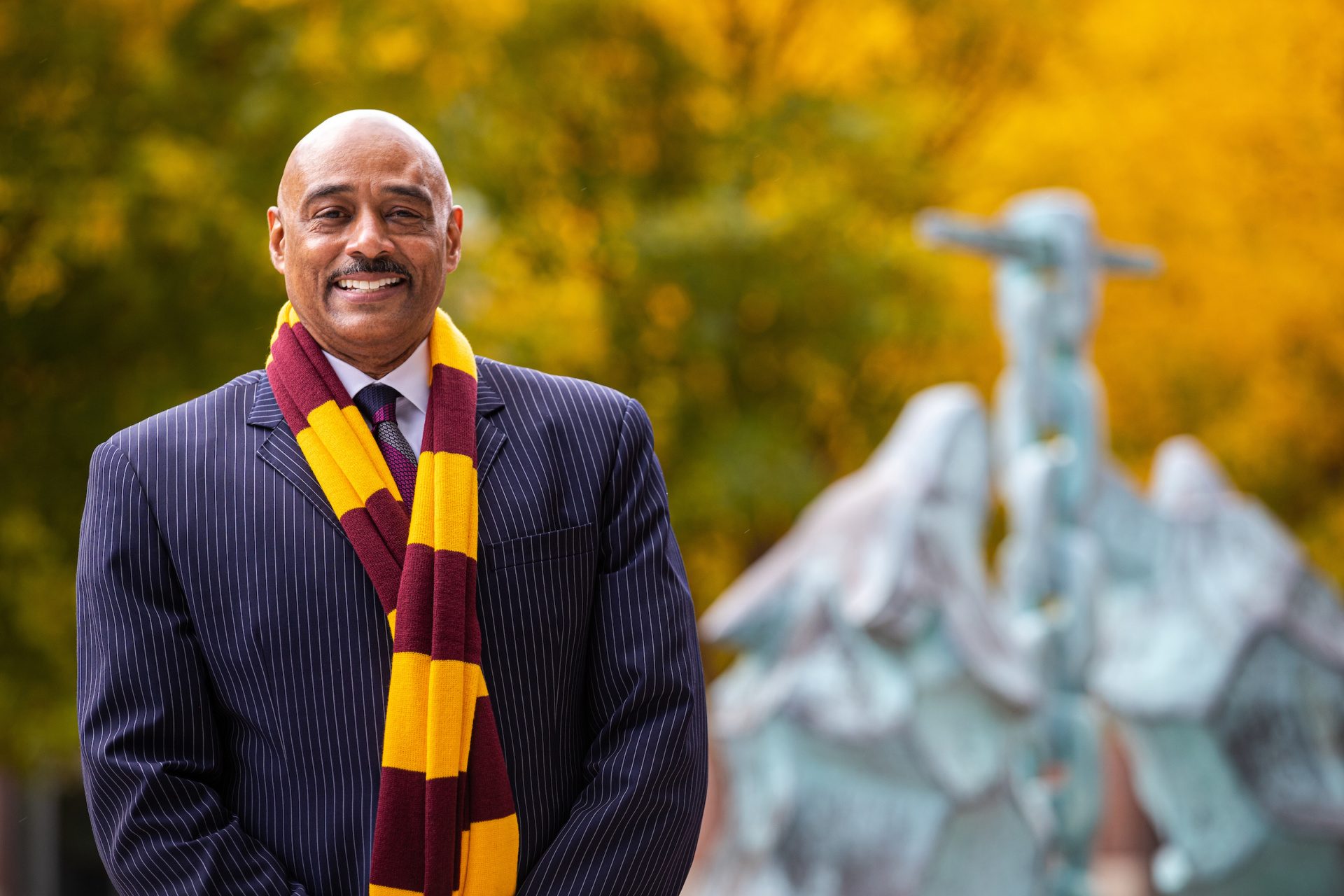Dr. Keith Champagne smiling wearing a Loyola maroon and gold striped scarf standing next to the Wolf and Kettle statue on Lake Shore Campus.