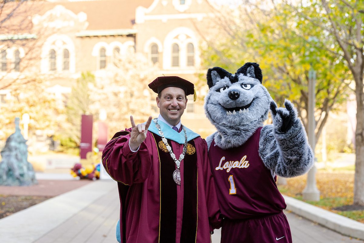 A man wearing maroon graduation regalia poses with a person wearing a wolf costume