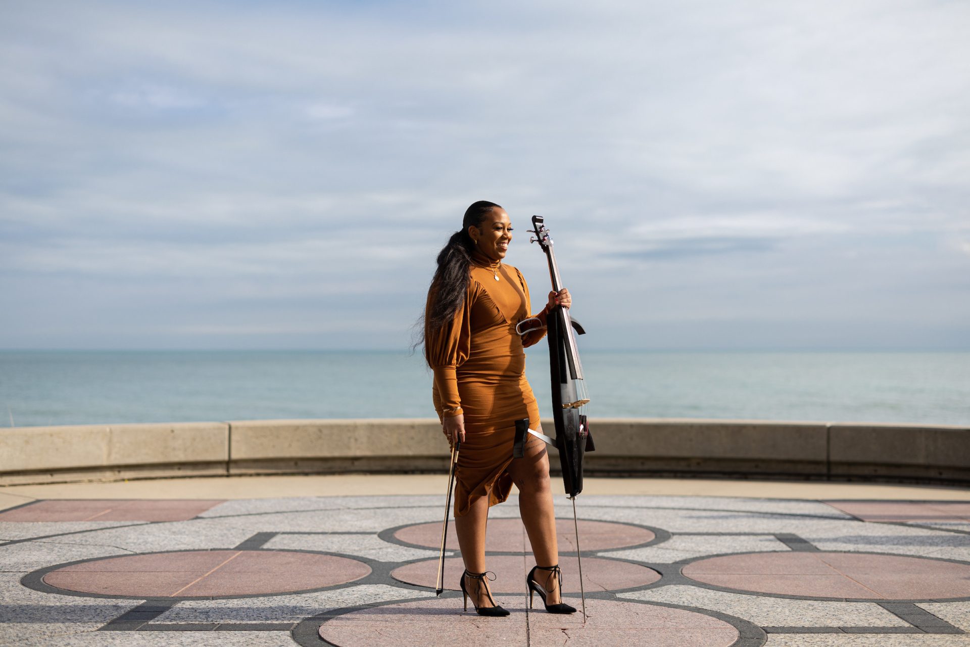 A Black woman stands on a brick sidewalk in front of a lake and holds a cello