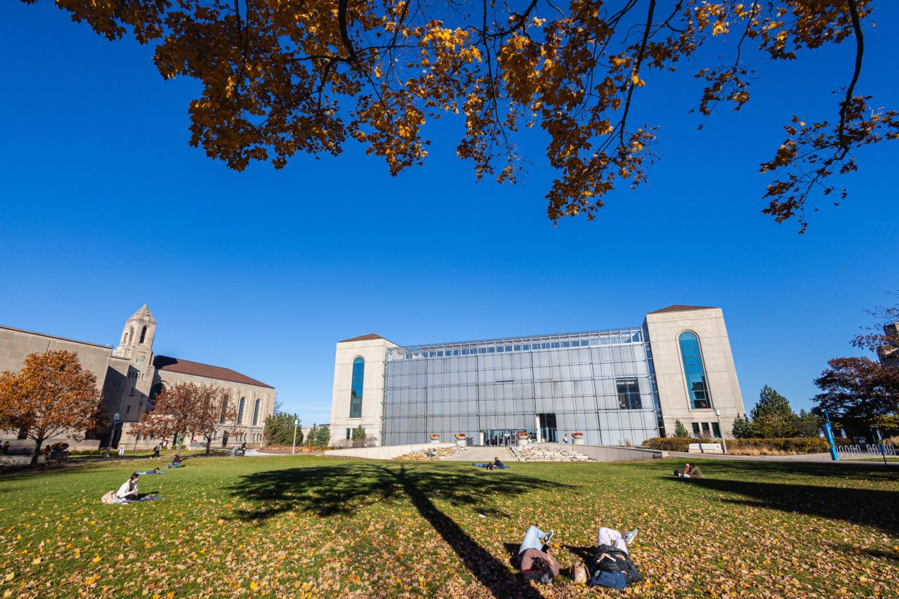 A Loyola University Chicago student relaxes outside a large glass building in grass littered with fall leaves