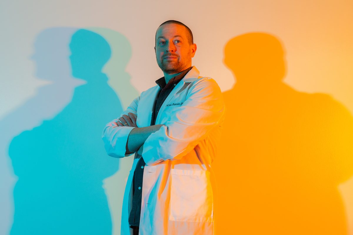 A man wearing a white lab coat stands in blue and orange lighting and crosses his arms