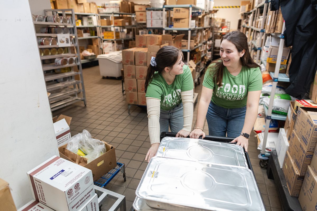 Two Loyola University Chicago students smile as they push a cart carrying foil food containers in an industrial kitchen
