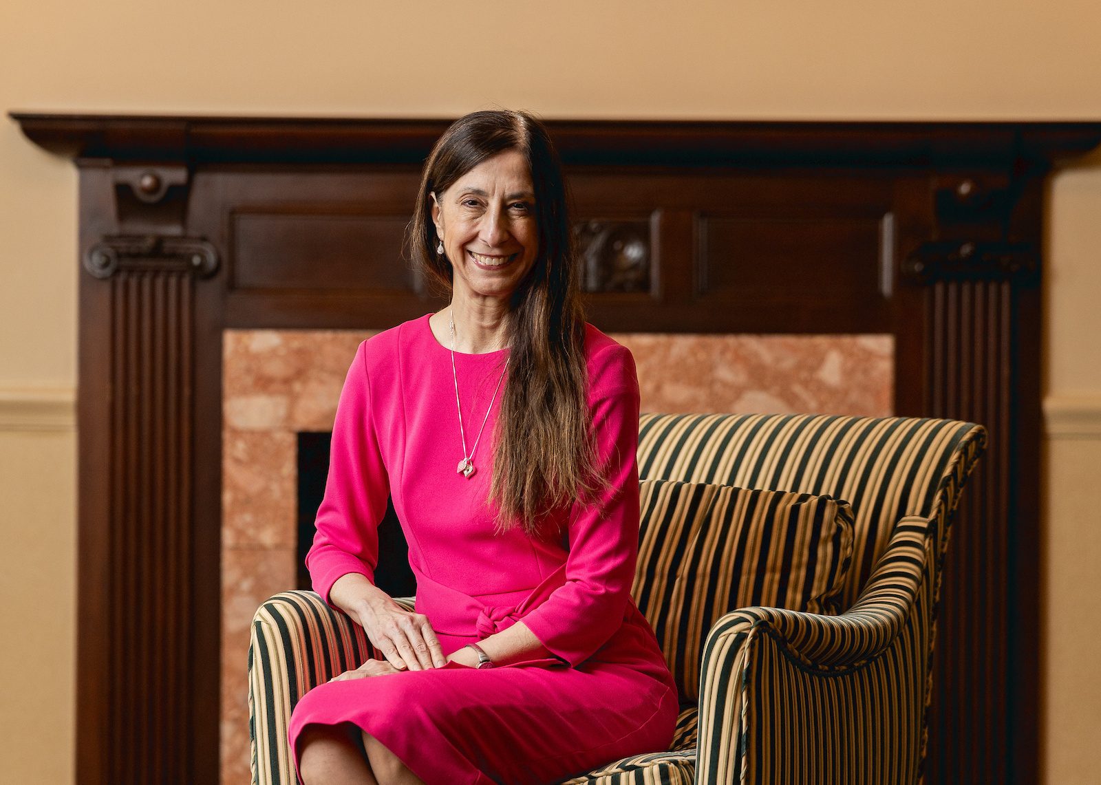 A woman in a hot pink dress smiles as she sits in a striped armchair