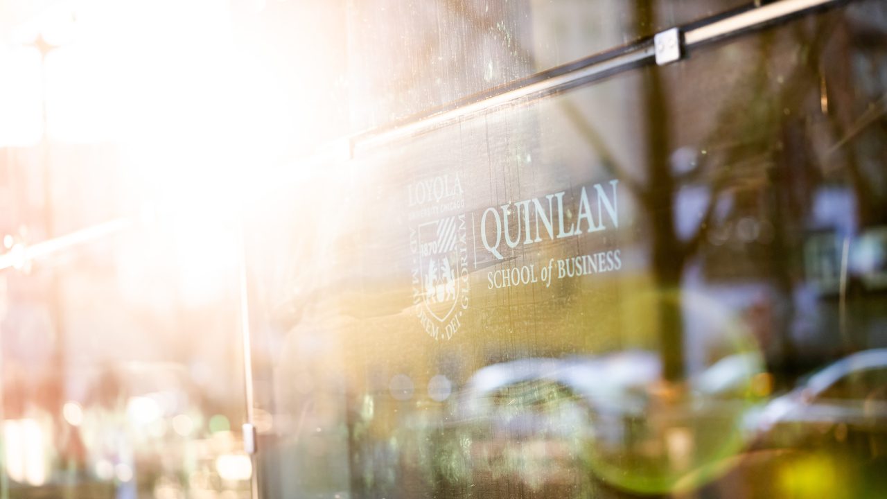 The sun shines upon a glass building that reads Quinlan School of Business