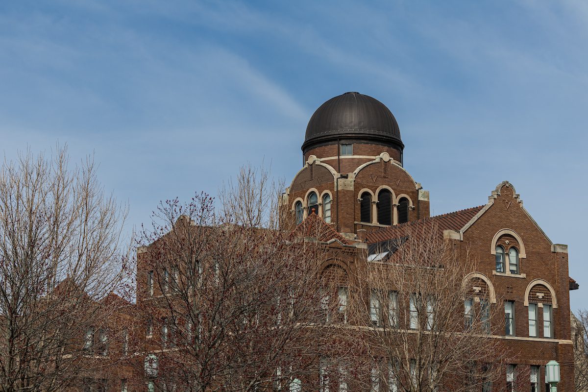 A red brick building with a large dome and white brick accents stands against a blue sky with leafless trees in the foreground
