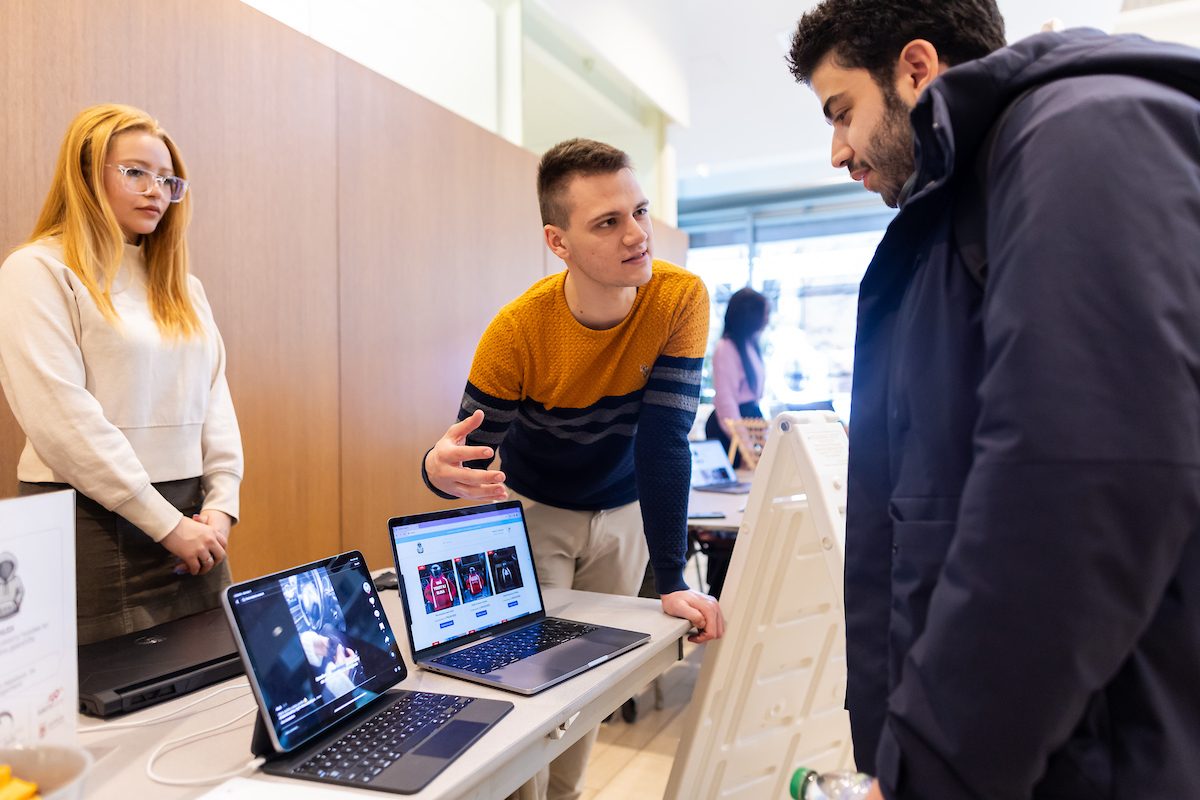 Quinlan School of Business students participate in the Side Hustle Showcase, designed to shine a spotlight on the innovative and enterprising endeavors of Loyola University Chicago students. Selected students showcase their entrepreneurial *side hustles* for judges to pick a winner. (Photo: Lukas Keapproth) A Loyola University Chicago students leans against a table with two laptops and explains what's on the screens