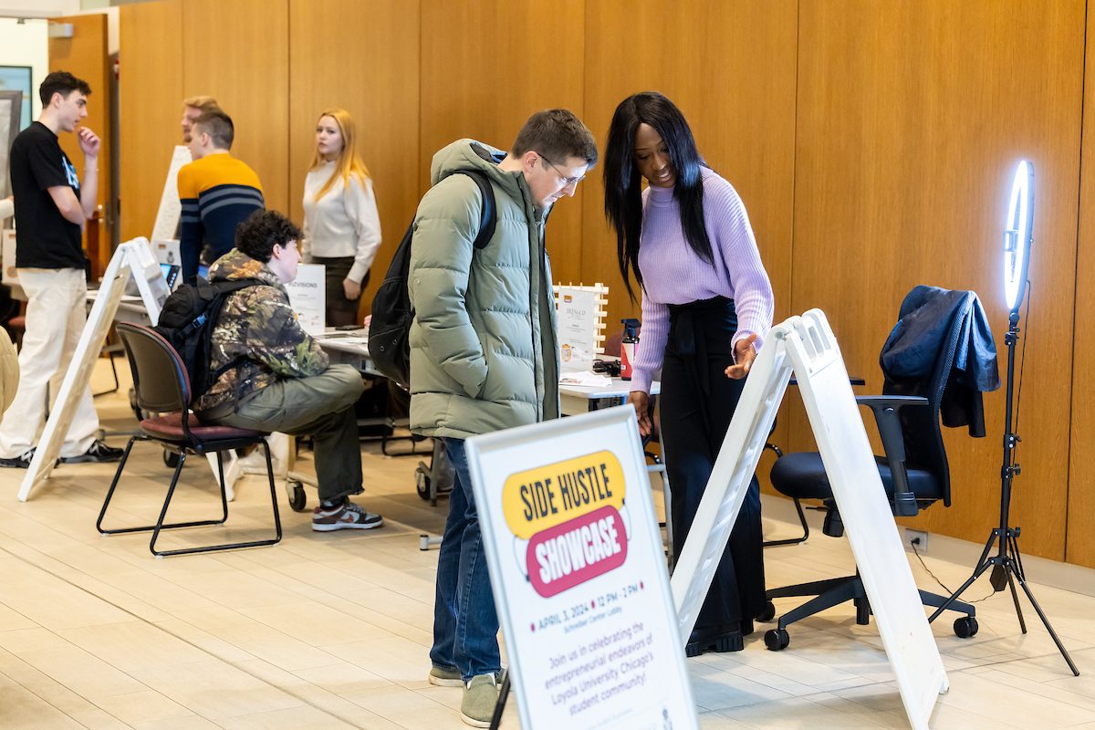 Quinlan School of Business students participate in the Side Hustle Showcase, designed to shine a spotlight on the innovative and enterprising endeavors of Loyola University Chicago students. Selected students showcase their entrepreneurial *side hustles* for judges to pick a winner. (Photo: Lukas Keapproth) A Loyola University Chicago student wearing a purple sweater gestures to a sign and speaks to a man wearing a green puffer jacket