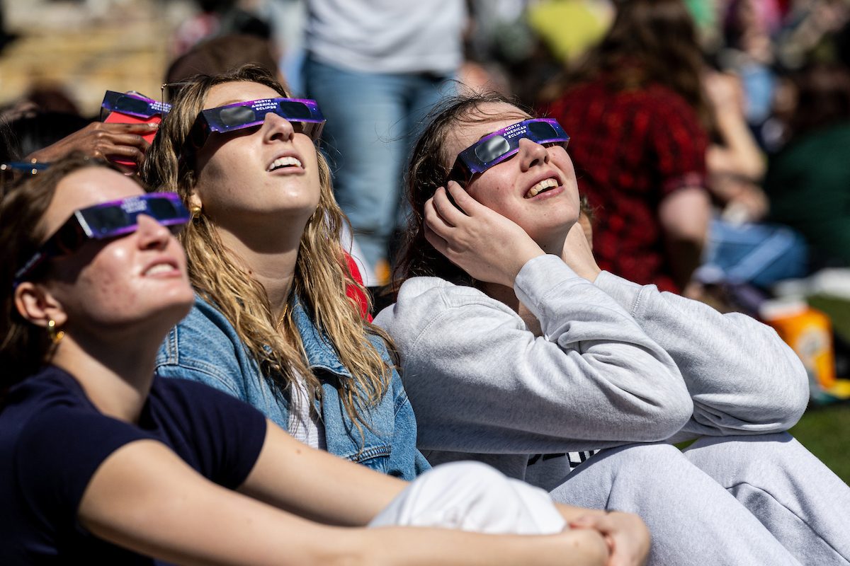 The Loyola University Chicago community gathered at the Lake Shore Campus to witness the historic, near total solar eclipse on April 8, 2024. (Photo: Lukas Keapproth) Three Loyola University Chicago students wearing solar eclipse glasses sit outdoors and look up toward the sky