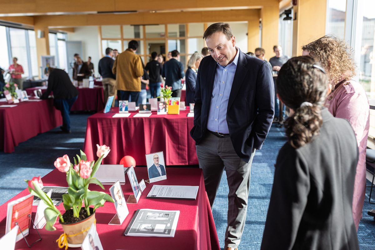 University Libraries hosts the annual Faculty Scholarship Celebration event in Information Commons on April 18, 2024. (Photo by: Lukas Keapproth) A man wearing a black blazer reads information on a rectangular table with a red tablecloth displaying books and author information as two women look on
