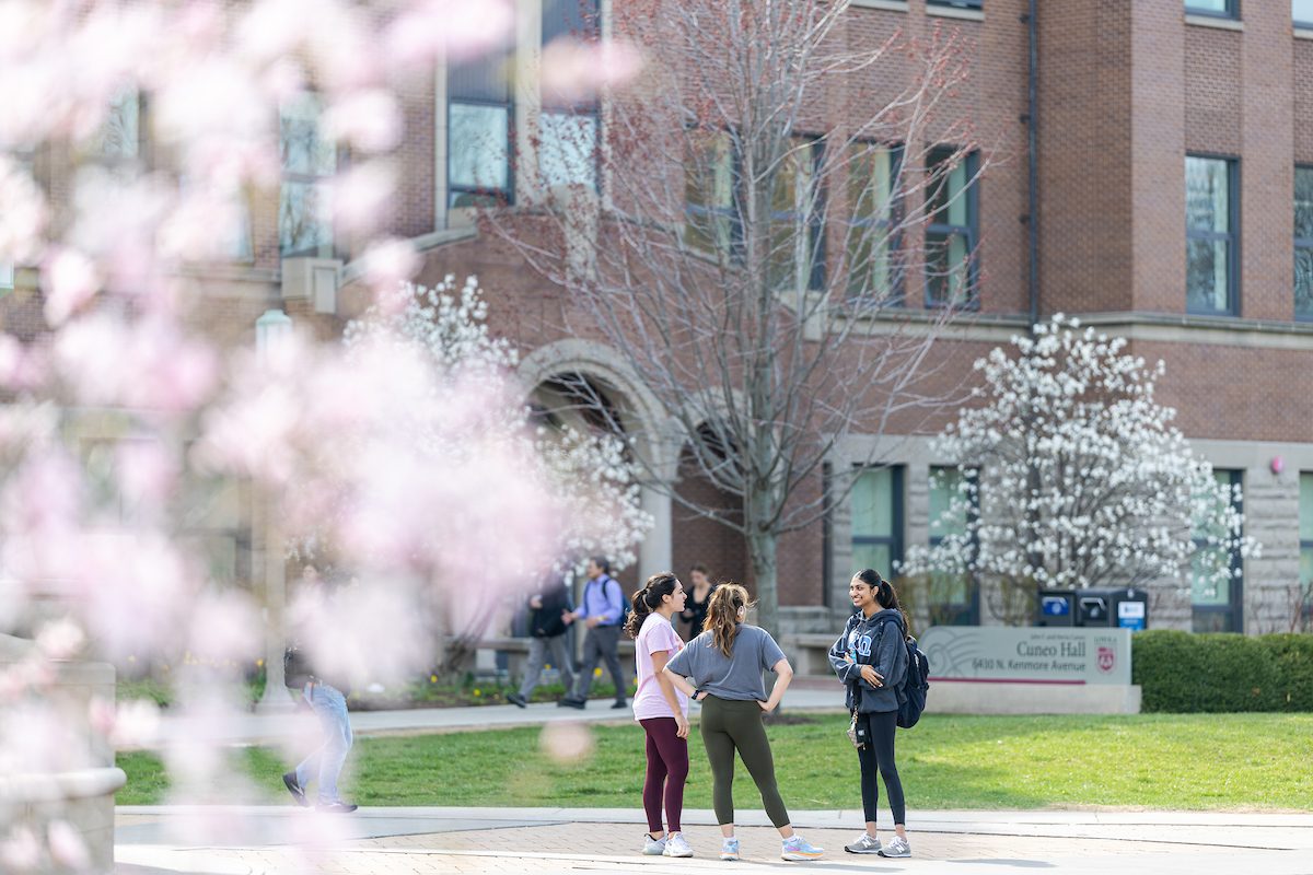 Spring weather on the Lake Shore Campus, April 10, 2024. (Photo by: Lukas Keapproth) Three Loyola University Chicago students stand outdoors and talk in front of a large red brick building with blooming trees planted in front