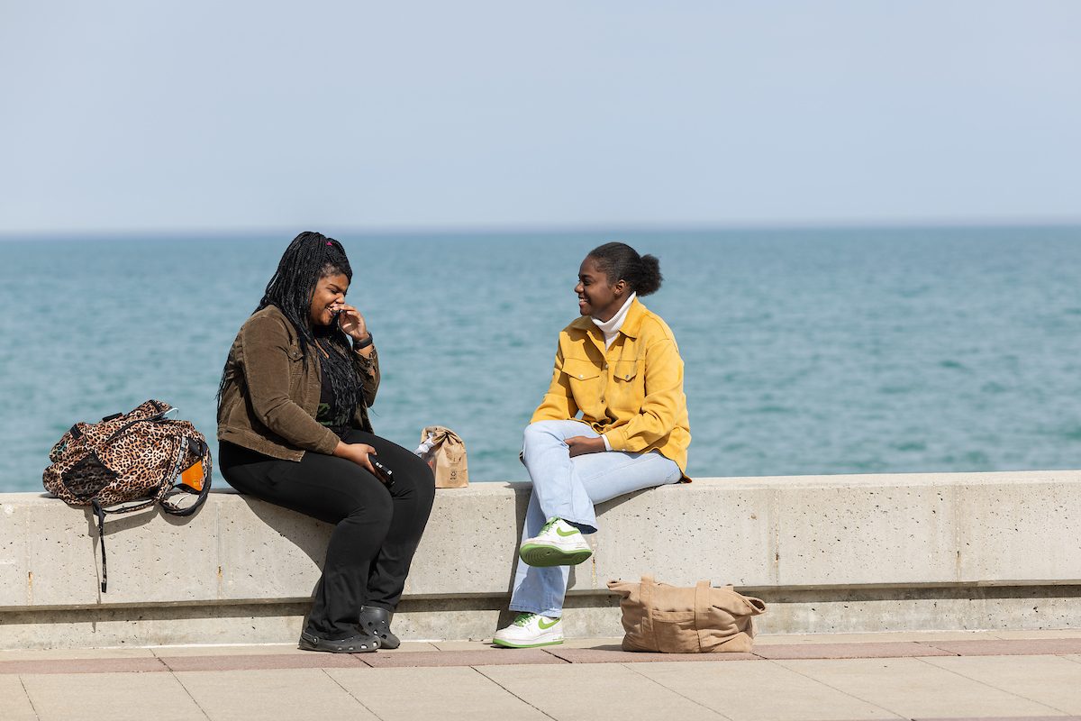 Spring weather on the Lake Shore Campus, April 10, 2024. (Photo by: Lukas Keapproth) Two Loyola University Chicago students sit and talk on a concrete bench in front of a large lake