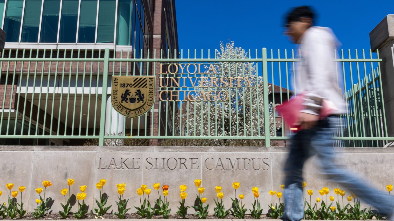 A person walks by a sign for Loyola University Chicago's Lake Shore Campus on a sunny, spring day.