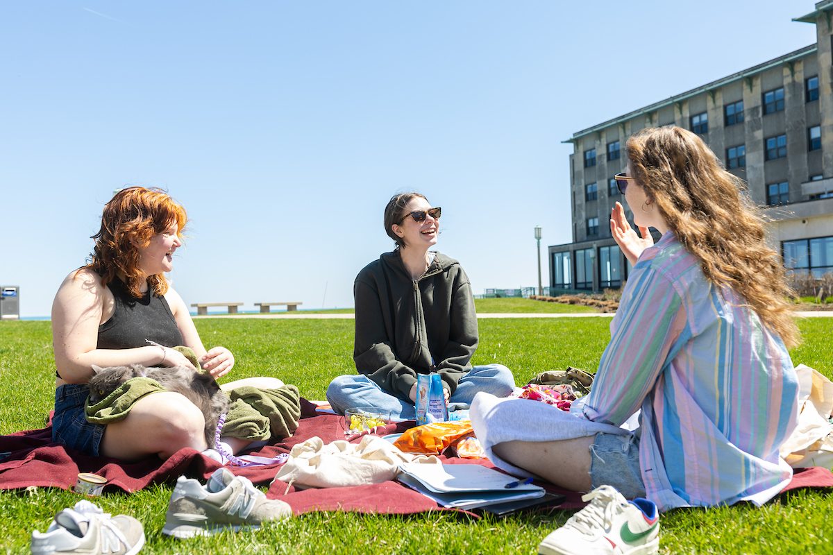 The Lake Shore Campus blooms with warmer spring weather, April 13, 2024. (Photo by: Lukas Keapproth) Three Loyola University Chicago students sit on a picnic blanket with snacks on a sunny day near a lake