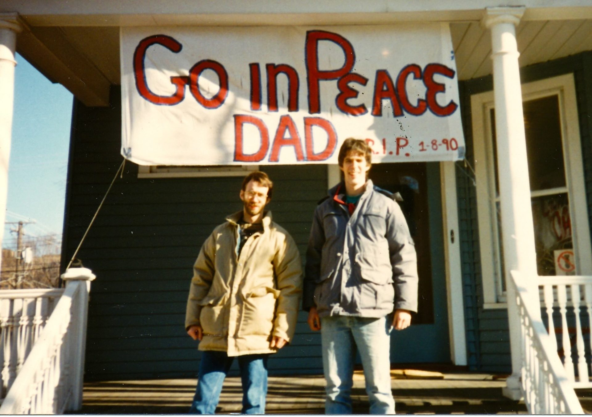 Two men wearing coats stand on a porch with white railing in front of a painted sign that reads 