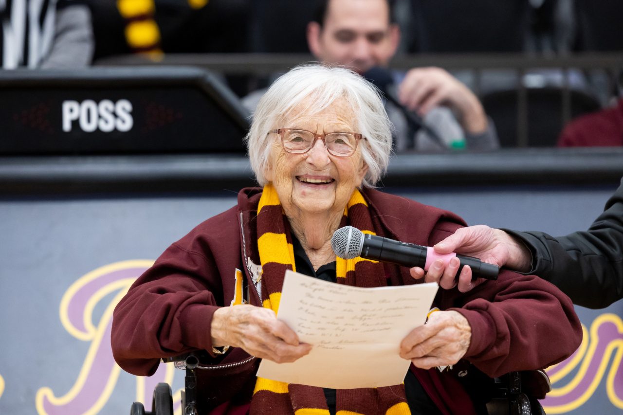 A woman in a wheelchair wearing a Loyola University Chicago scarf holds a sheet of paper and speaks into a microphone in a sports stadium