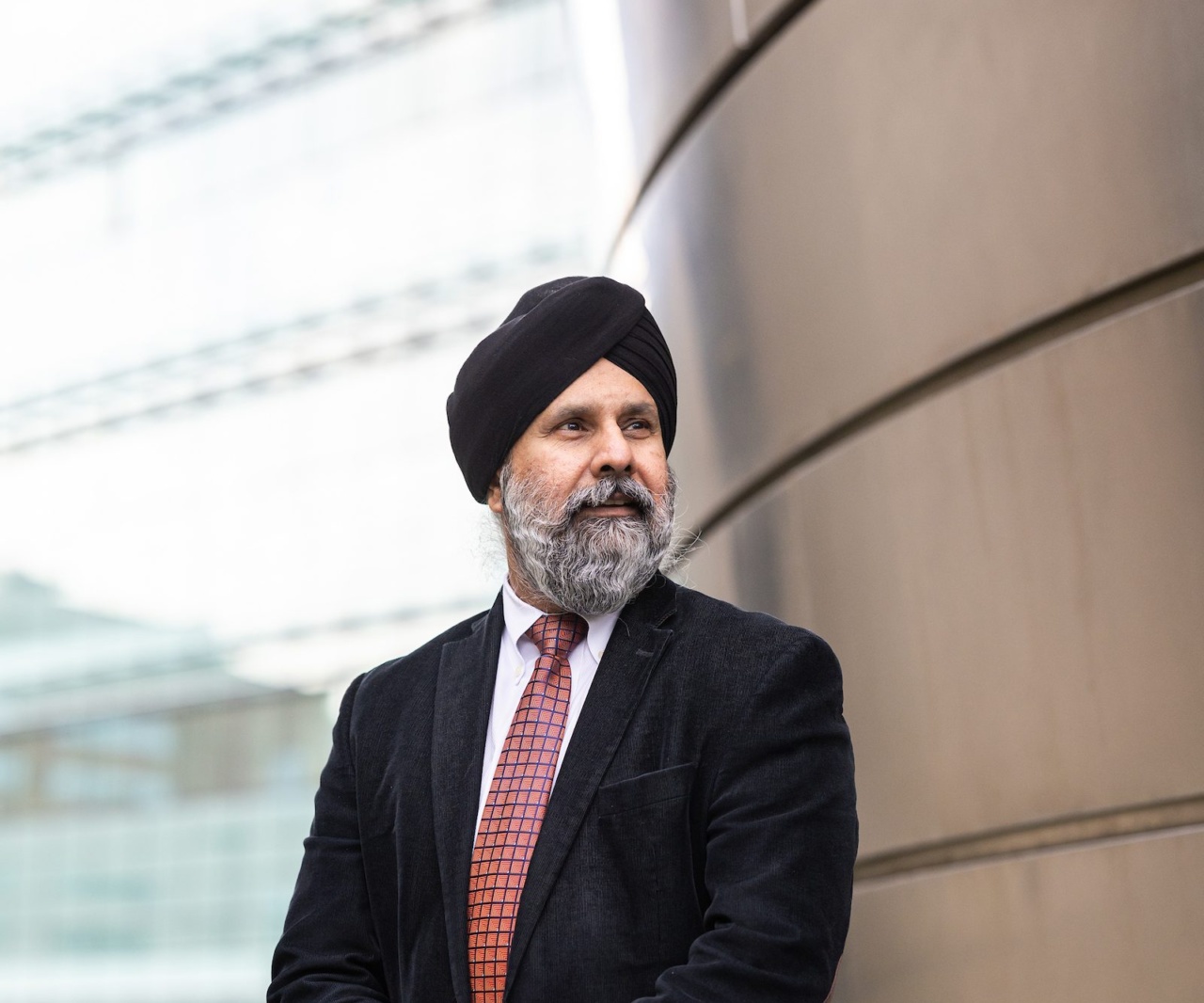 A man wearing a black turban, a black suit, and a red tie stands outdoors with his hands clasped on the Loyola University Chicago campus