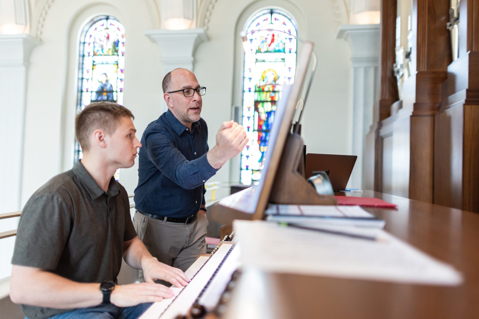 A man instructs a Loyola University Chicago student on the organ with stained glass windows in the background