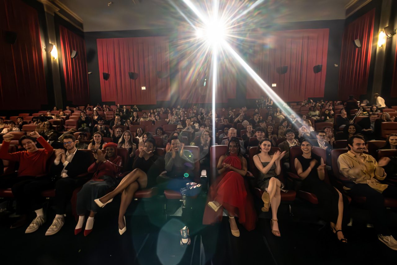 Loyola University Chicago students in formal wear sit in theater seats in a theater with red curtains as a spotlight shines at the camera