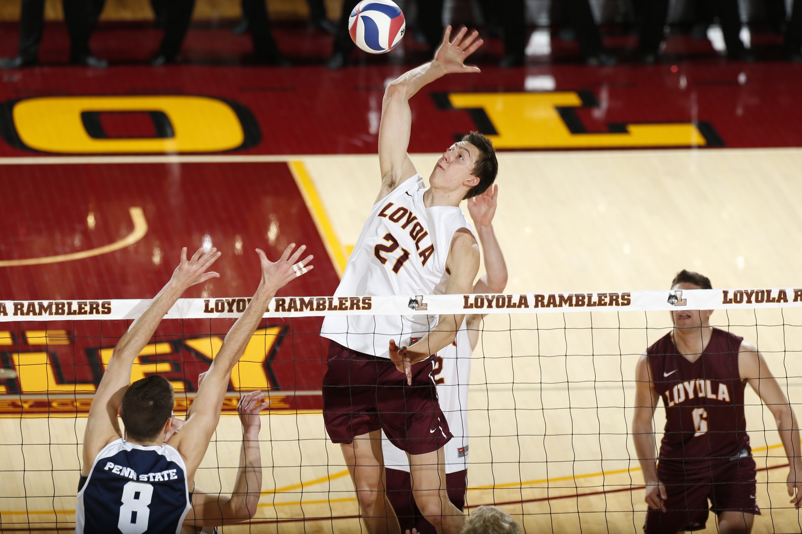 A man in a Loyola University Chicago jersey jumps in midair and prepares to spike a volleyball in front of a net as an opponent blocks on the other side on an indoor court with maroon and gold paint
