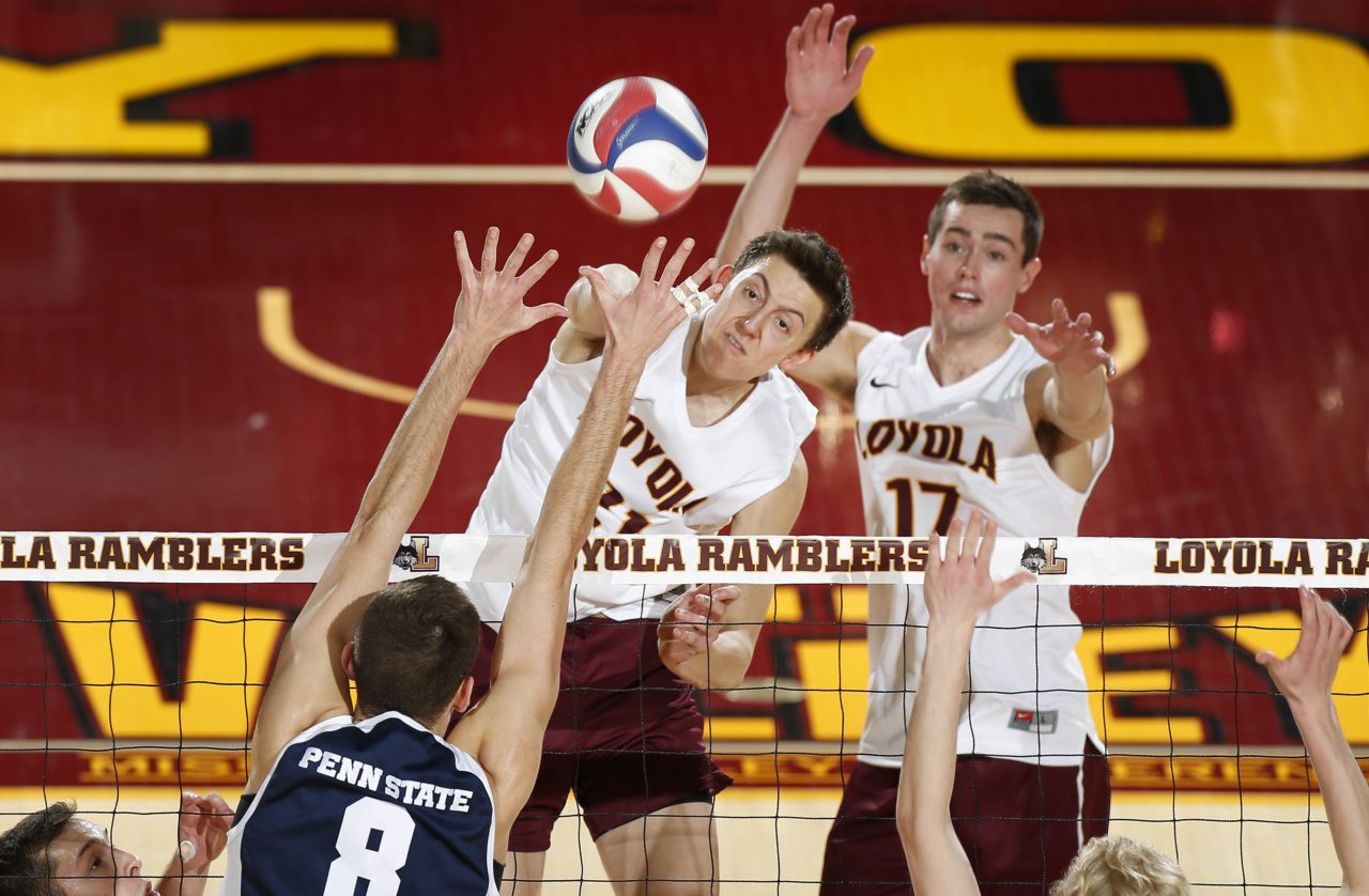 Two men in Loyola University Chicago jerseys jump to spike a ball as an opponent blocks on an indoor court with maroon and gold paint