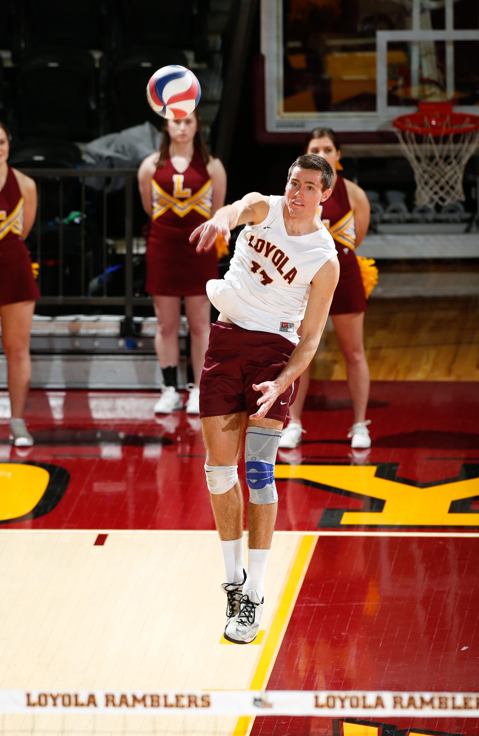 A man in a Loyola University Chicago jersey serves a volleyball on an indoor court with maroon and gold paint