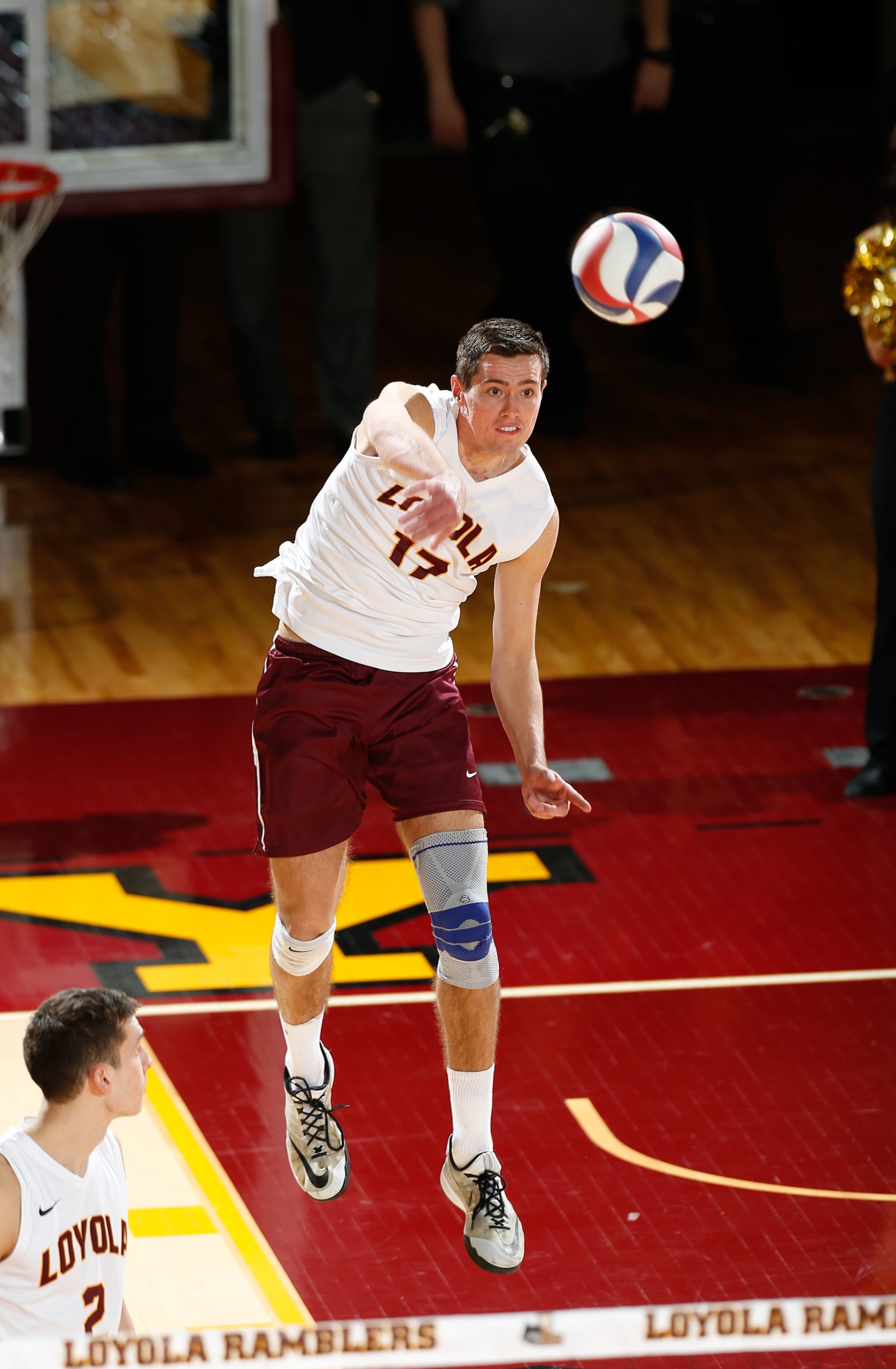A man wearing a Loyola University Chicago jersey jumps in midair and serves a volleyball on an indoor court with maroon and gold paint