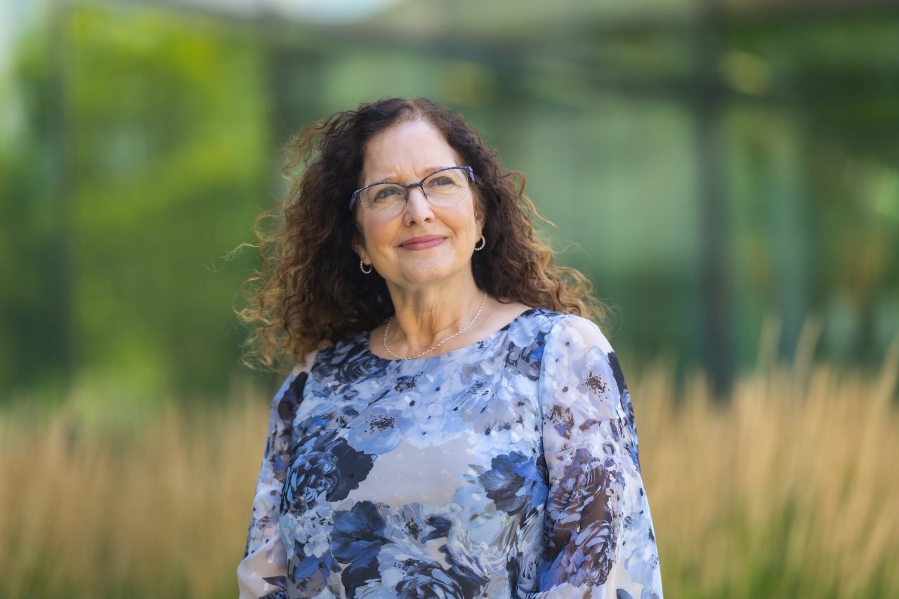 A woman wearing a blue floral shirt looks into the distance with greenery in the background