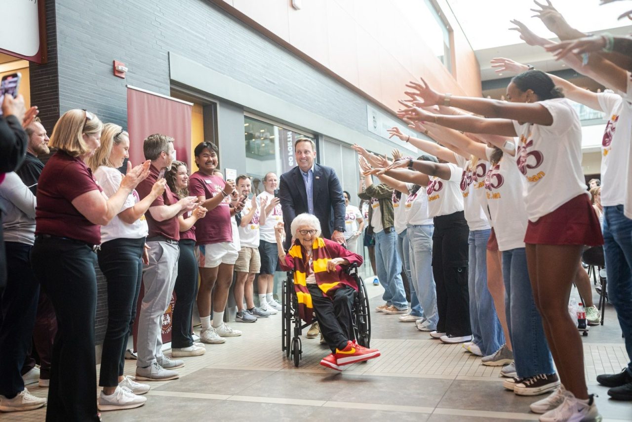 A man in a suit pushes an older woman in a wheelchair as Loyola University Chicago students hold up their hands in a cheer tunnel