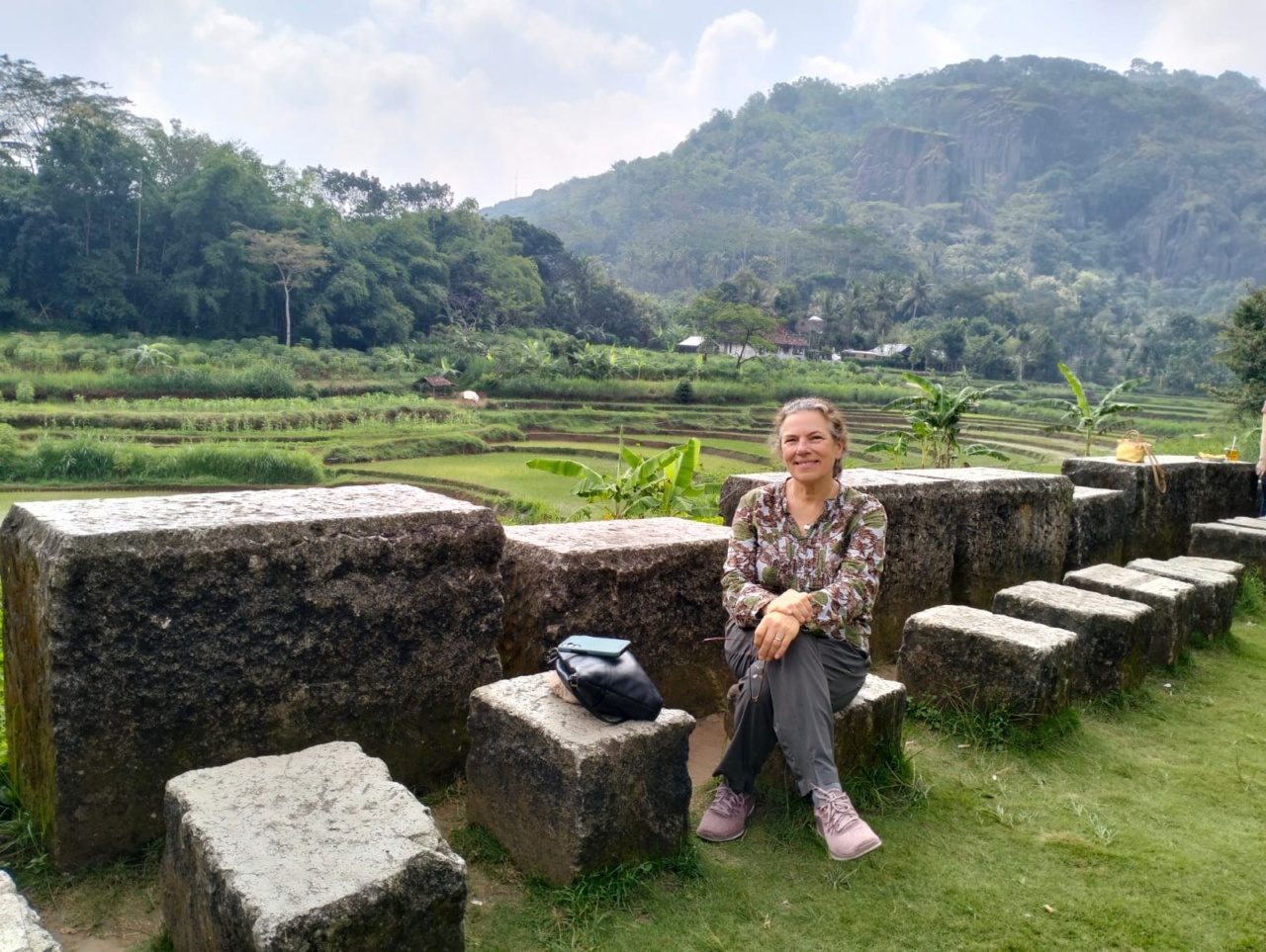 A Loyola University Chicago professor sits on a stone formation with foothills in the background in a lush landscape