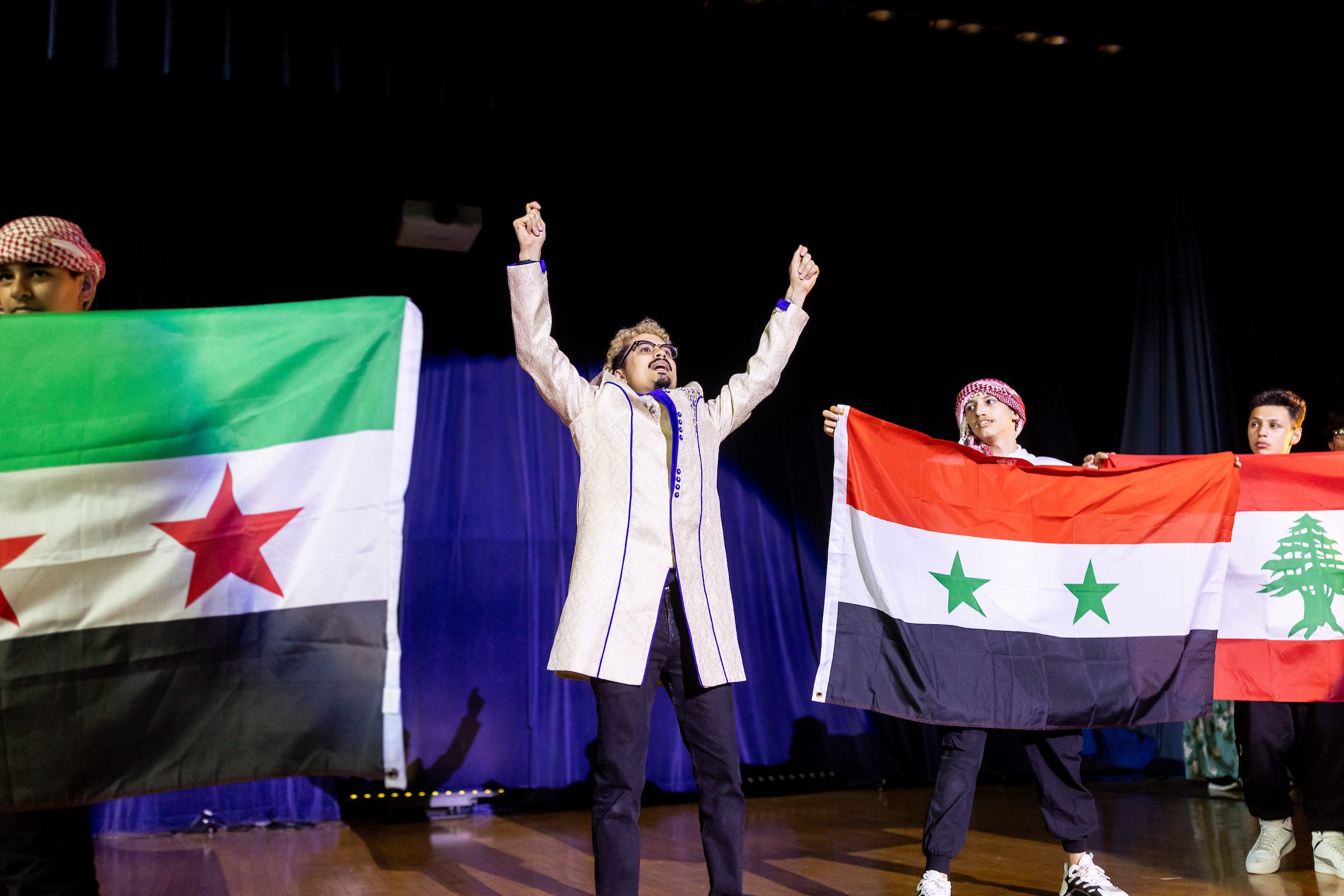 A Loyola University Chicago alum stands on a stage with his hands up between high school students holding flags for different countries