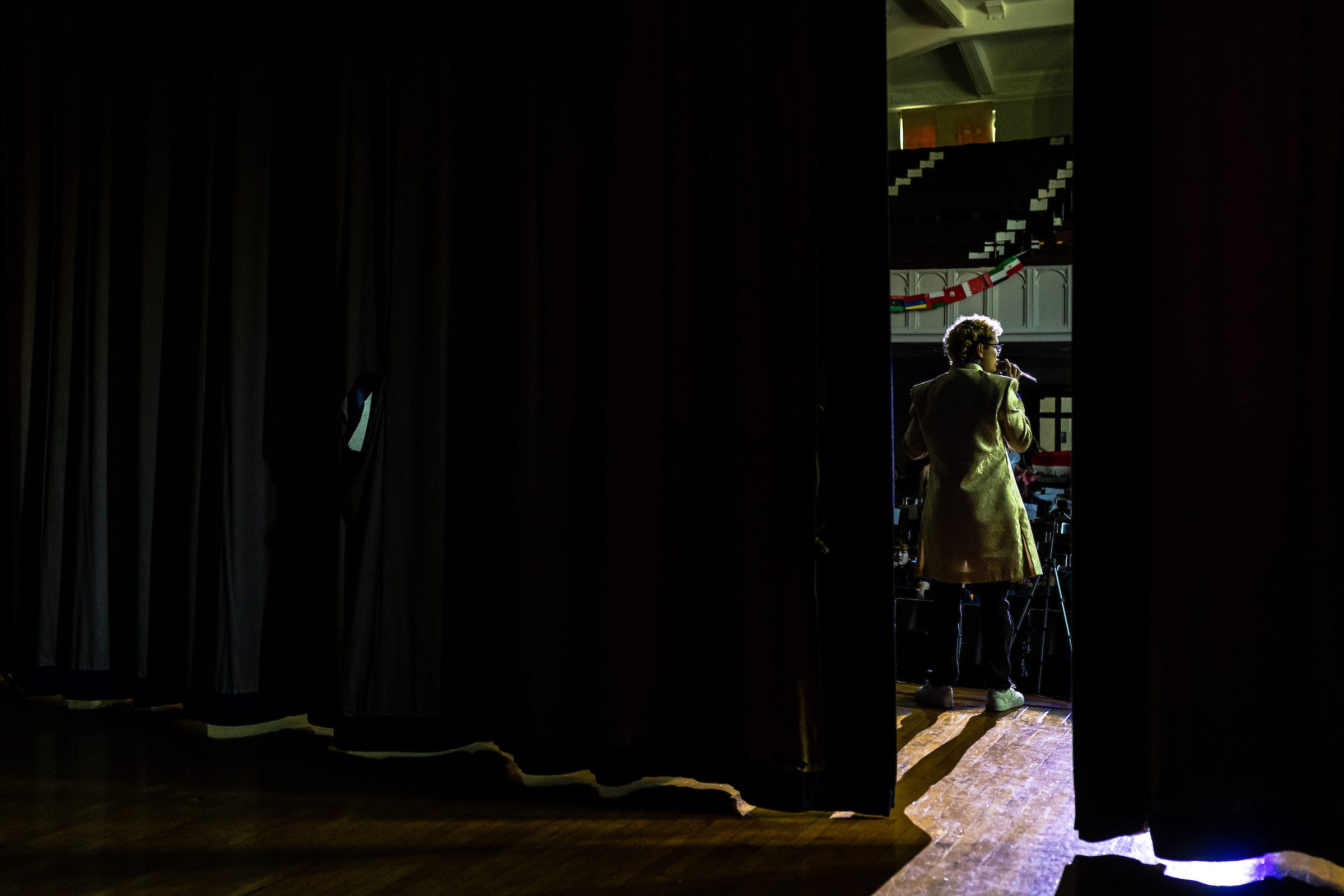A Loyola University Chicago alum and social worker at Sullivan High School speaks into a microphone on a stage, as seen through a gap in the stage's curtains