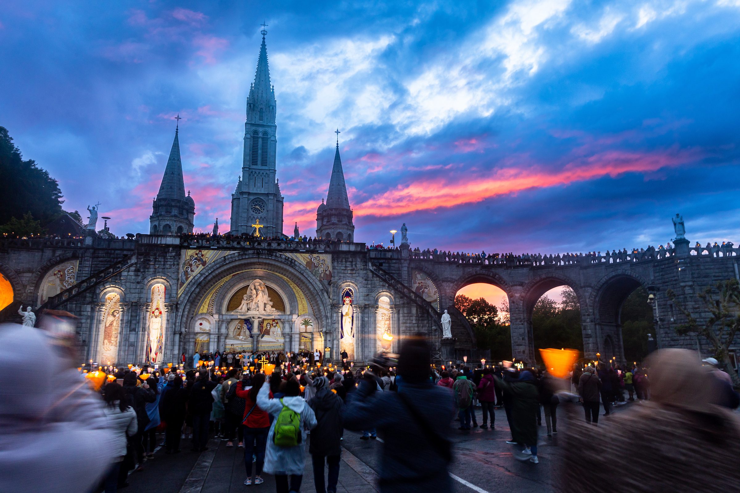 A crowd gathers in front of a stone building with tall towers at sunset
