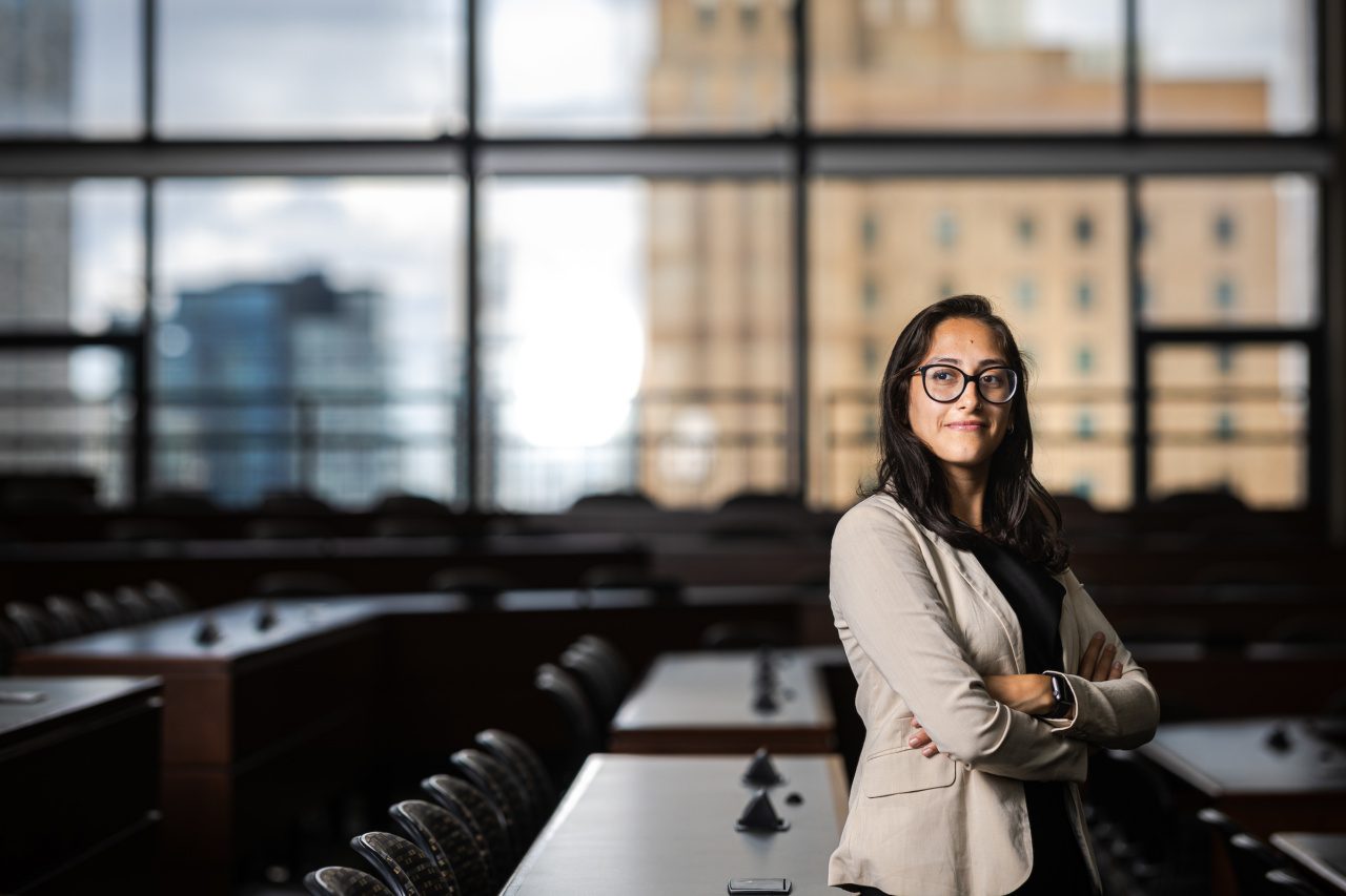 A Loyola University Chicago student stands with her arms crossed in a lecture hall with a wall of windows in the background showing buildings in downtown Chicago