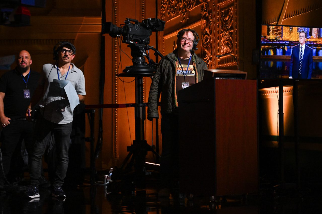 A group of men wearing headsets on a film set with cameras, lights, and a television screen showing Stephen Colbert