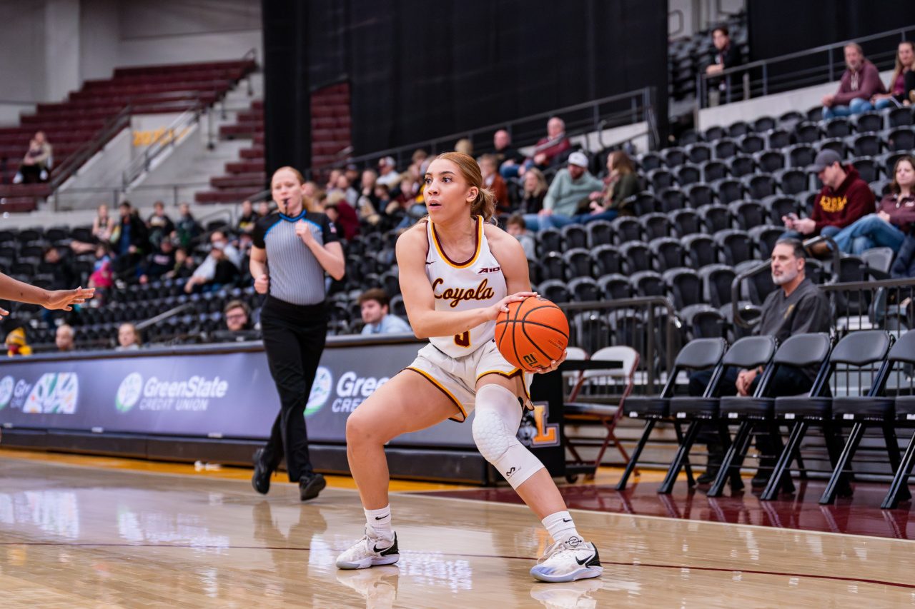 A Loyola University Chicago student wearing a basketball uniform prepares to pass a basketball to one of her teammates in a basketball arena during a game