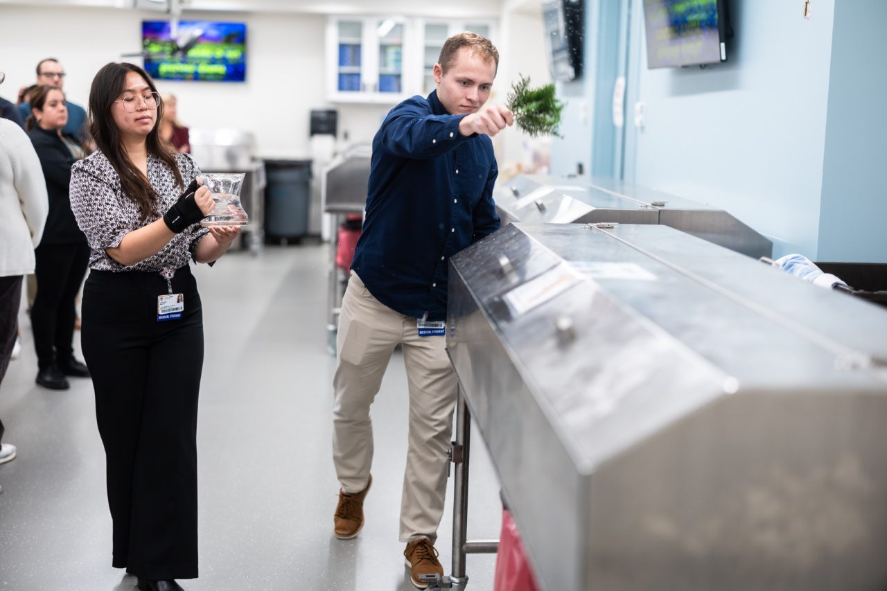 A Loyola University Chicago student performs a blessing over a metal case with a branch as another student holds a cup of water nearby