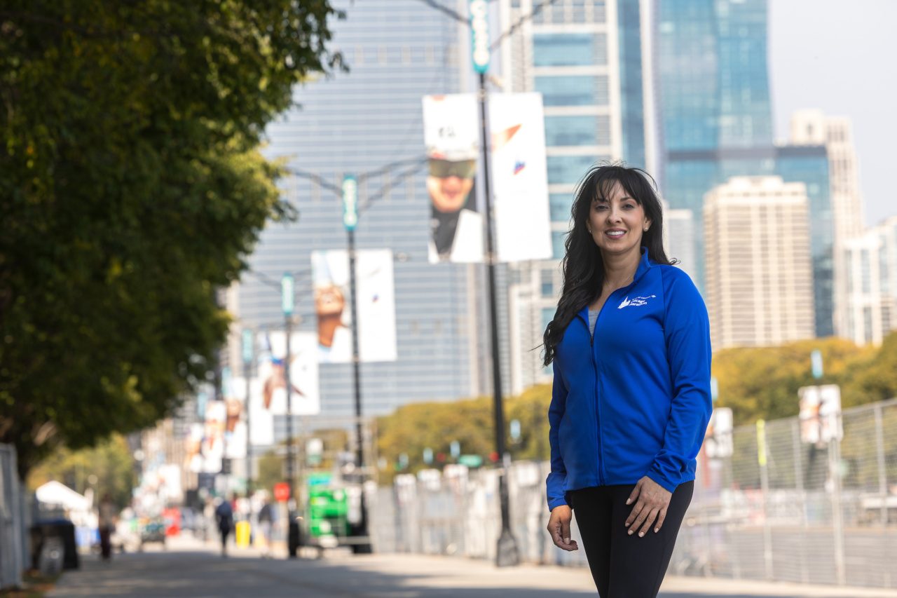 A woman wearing a blue jacket stands in front of a street with Chicago Marathon signage on lightposts and the downtown Chicago skyline in the background