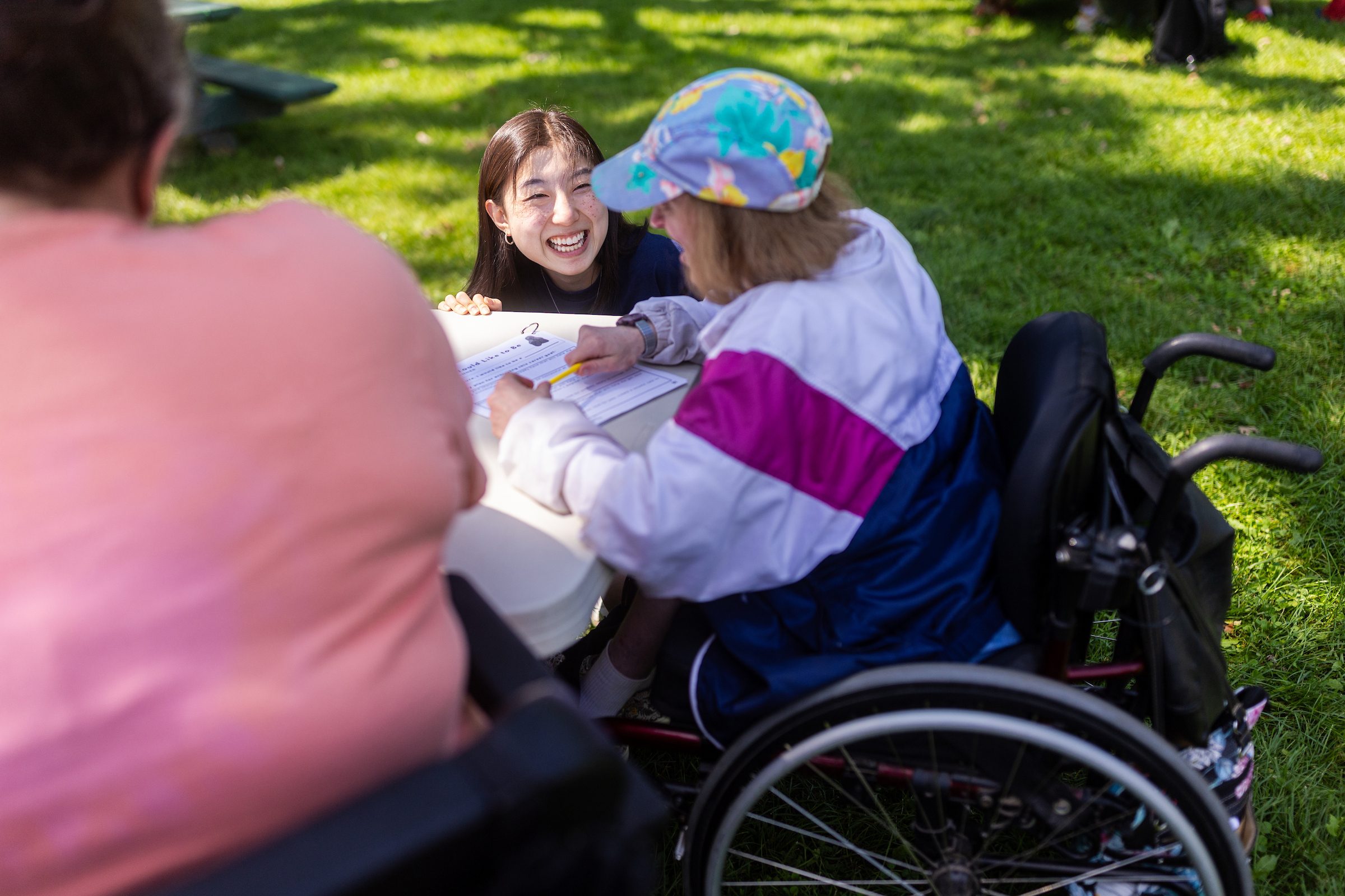 A woman smiles as she speaks to a woman using a wheelchair sitting outdoors at a white picnic table