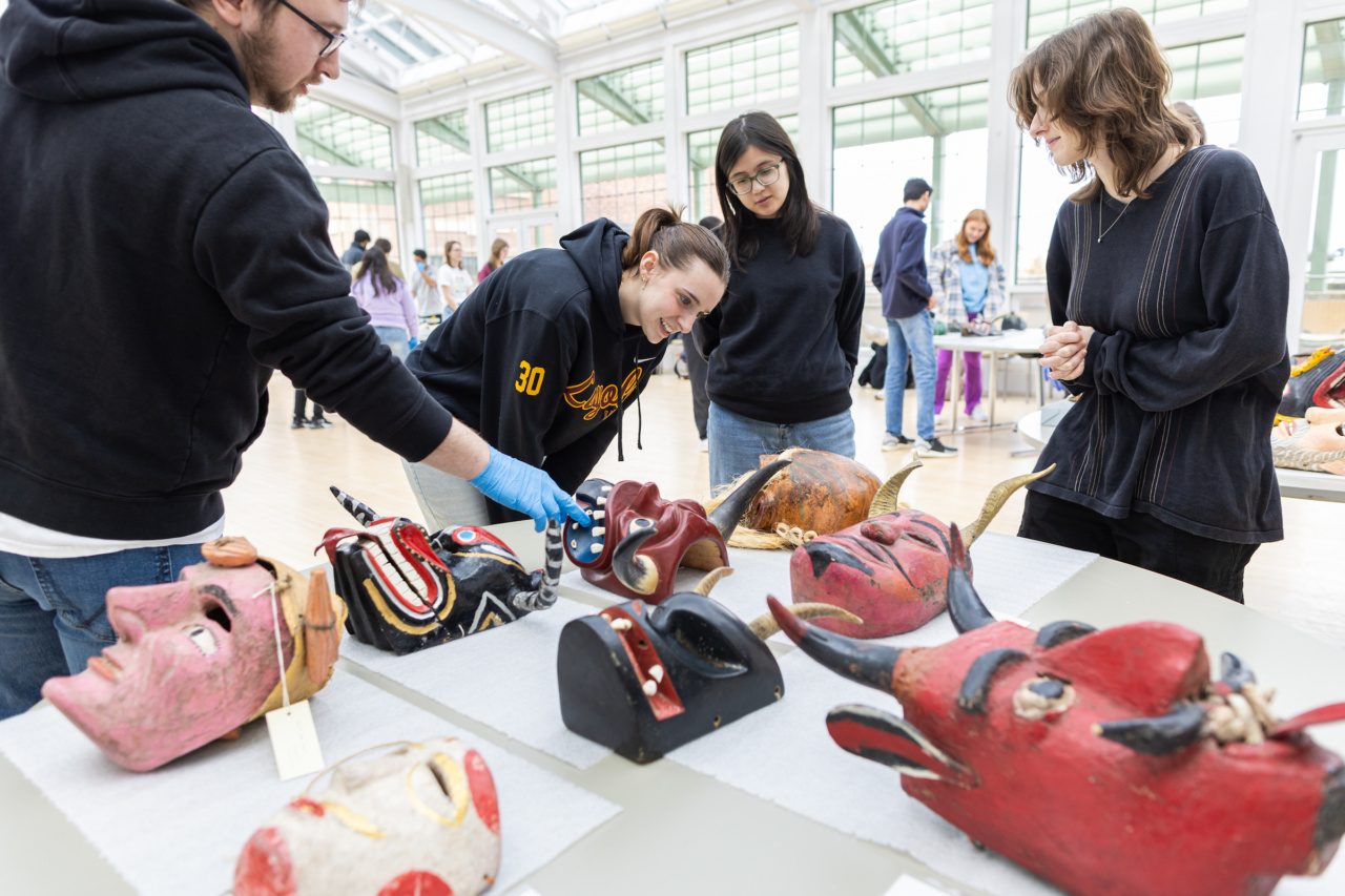 Loyola University Chicago students inspect tribal masks lying on a table in a large room with floor-to-ceiling windows